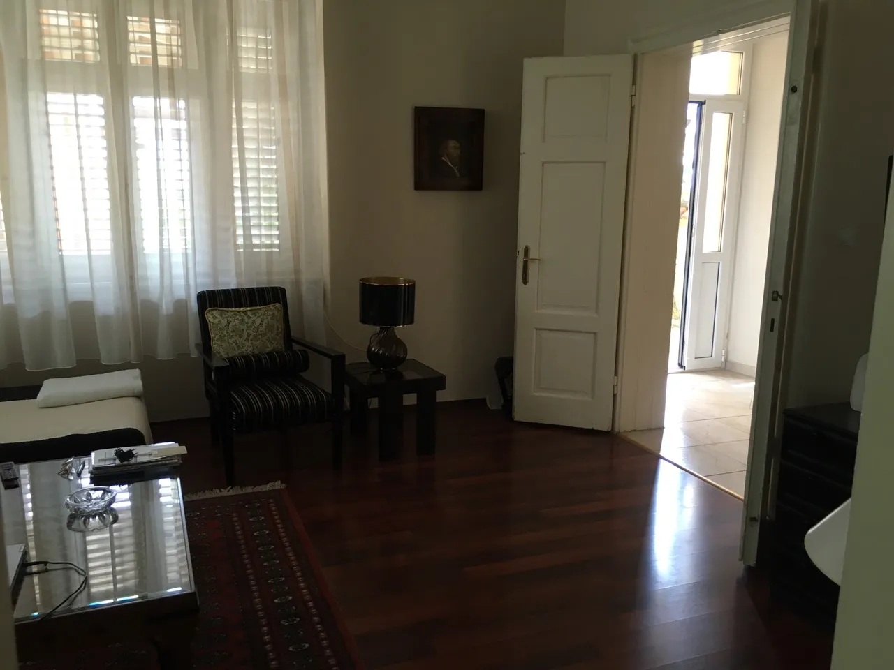 Living room with dark wood floors, striped armchair, glass coffee table, sheer curtains, and portrait on wall.