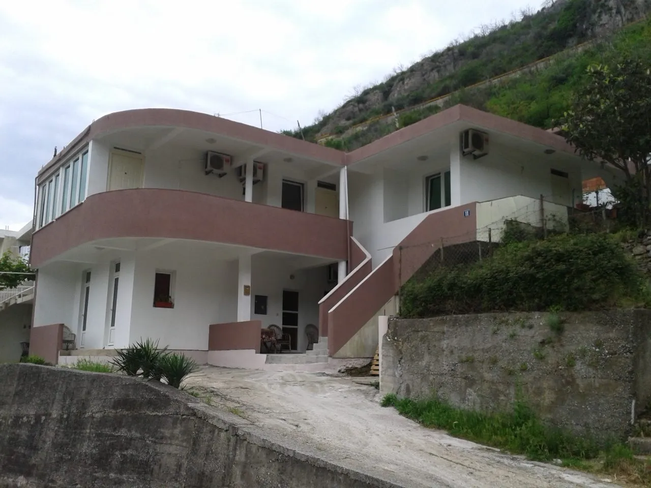 Exterior view of a two-story house with a terrace and AC units against a hillside.
