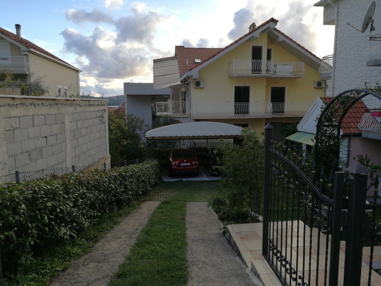 Two-story house in Tivat, Montenegro, with private covered parking, a green yard, and wrought-iron gates.