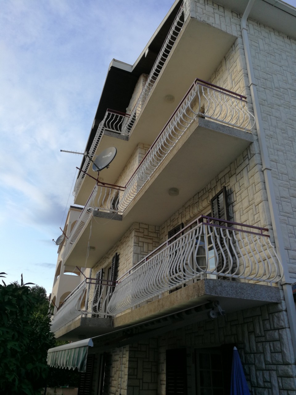 Facade of a three-story house in Tivat, Montenegro, with stone cladding and wrought-iron balconies on each floor.