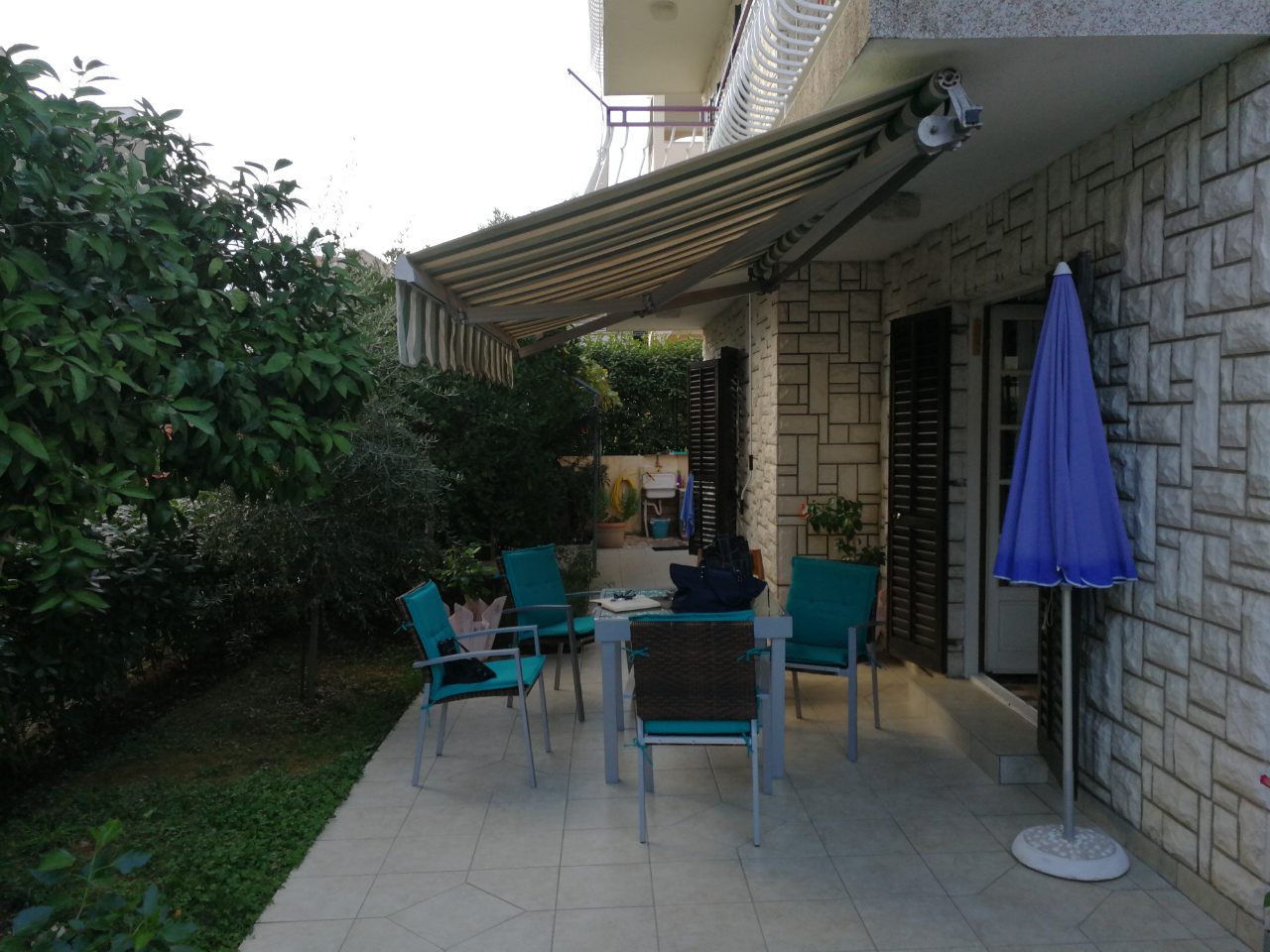 Cozy terrace with garden furniture and an awning by the house in Tivat, Montenegro, with a stone-clad wall and a green garden.