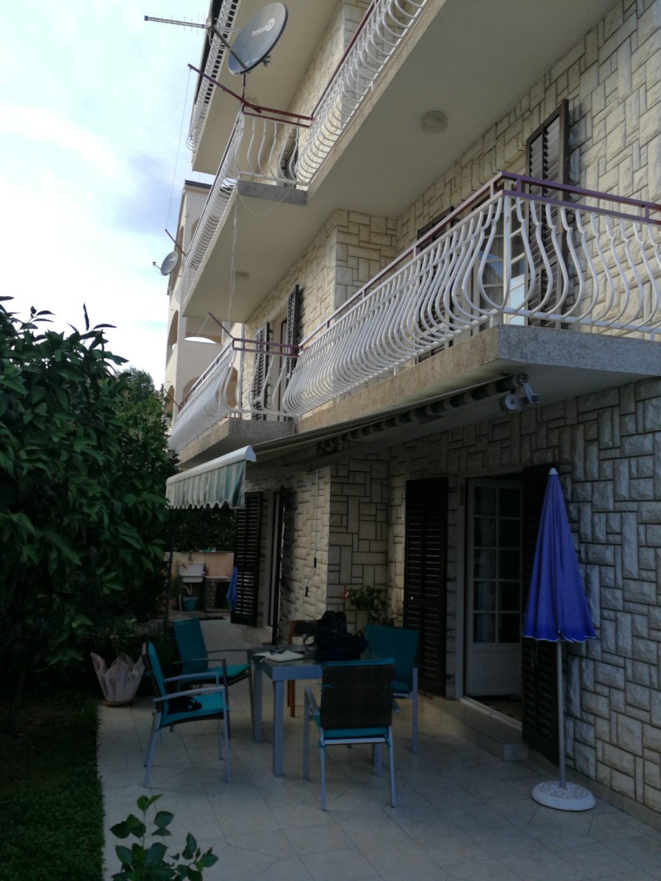 House facade in Tivat, Montenegro: stone cladding, balconies with wrought-iron railings, and a terrace with lounge furniture.