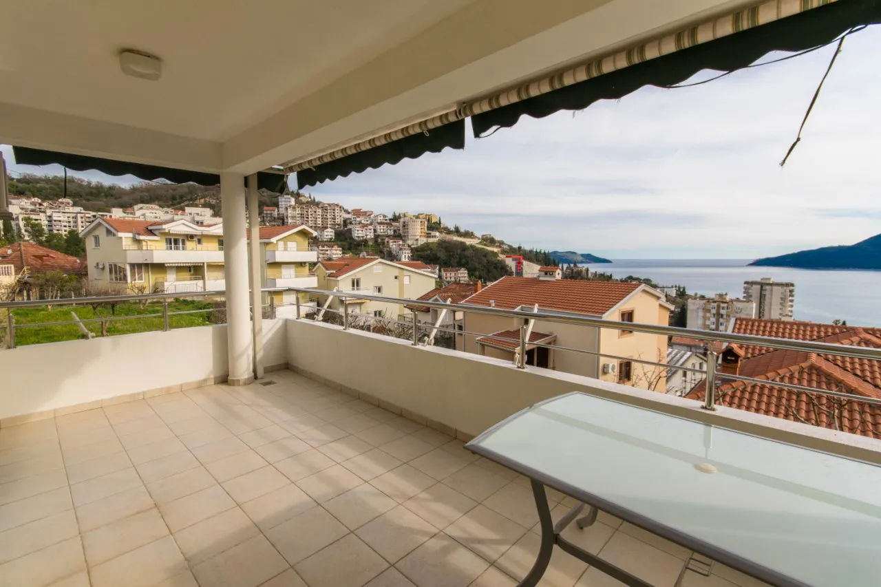 Tiled terrace with a glass table offering a scenic view of coastal houses and the sea.