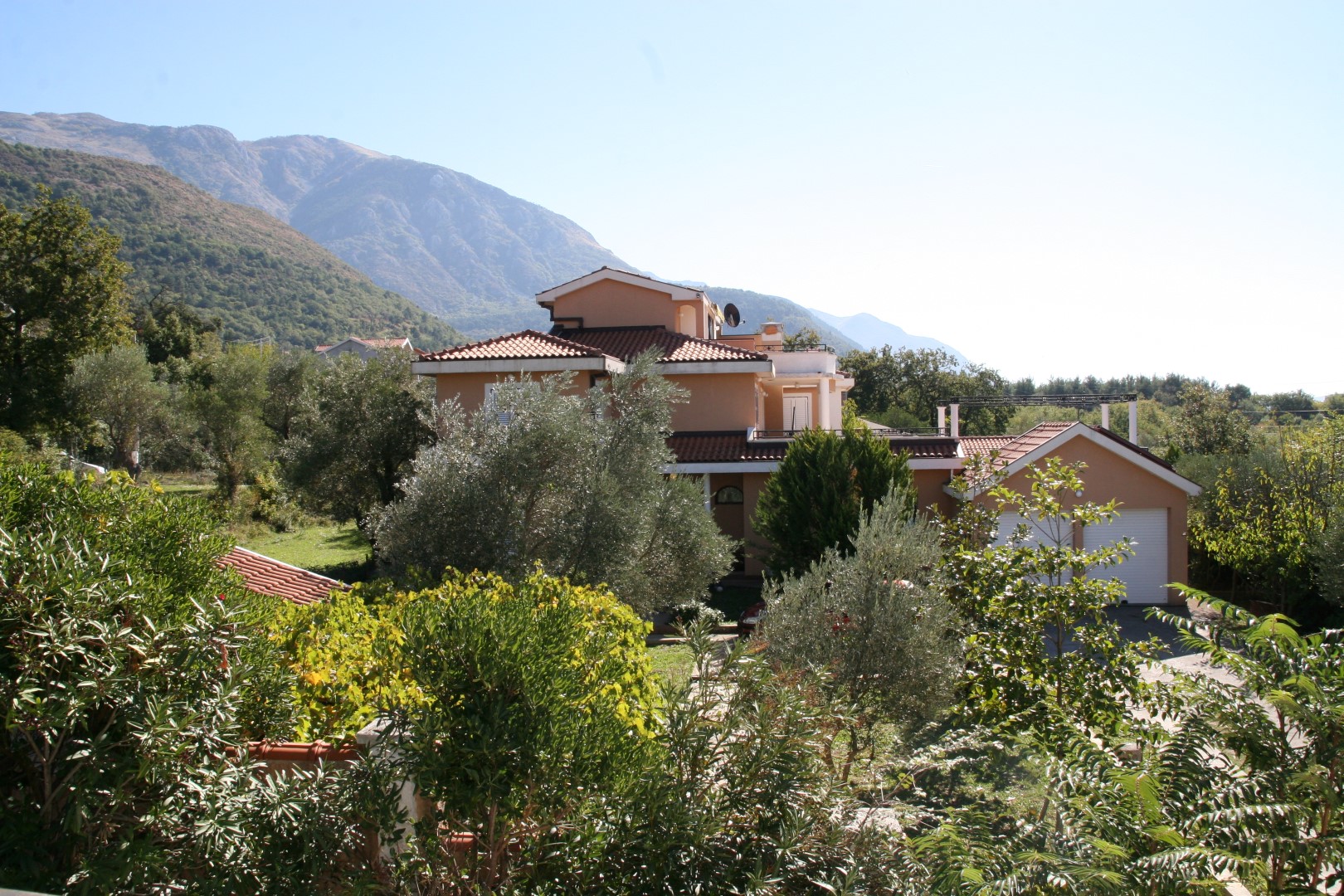 Two-story villa with a garage in Kavači, Montenegro, with mountains in the Bay of Kotor in the background, surrounded by greenery and olive trees.