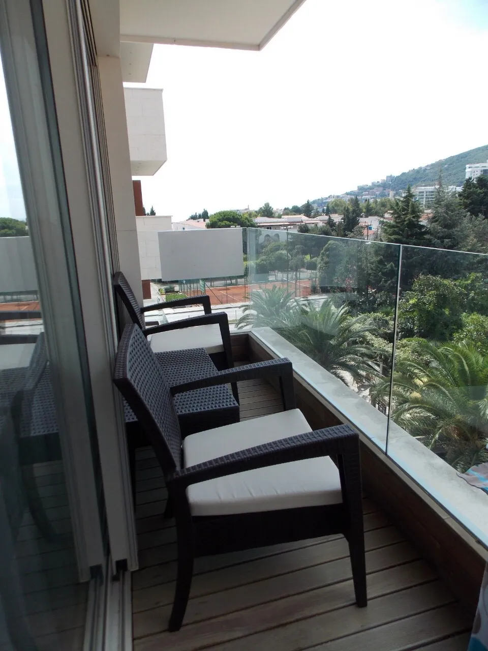 Balcony with dark wicker chairs and white cushions, wooden decking, and a glass railing overlooking trees.