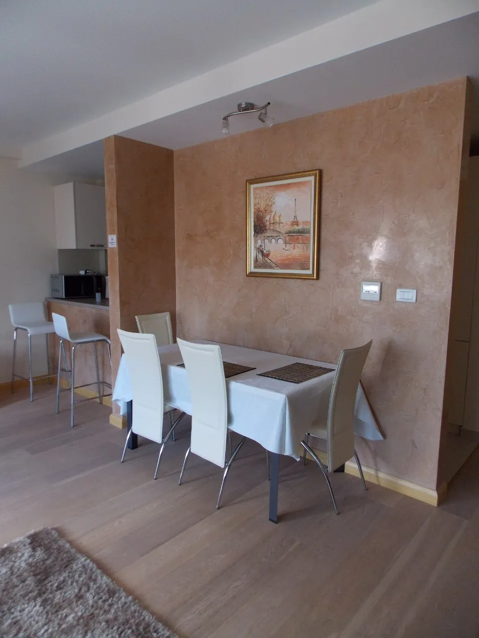 Dining area with white table and chairs against a beige wall; partial kitchen visible on the left.