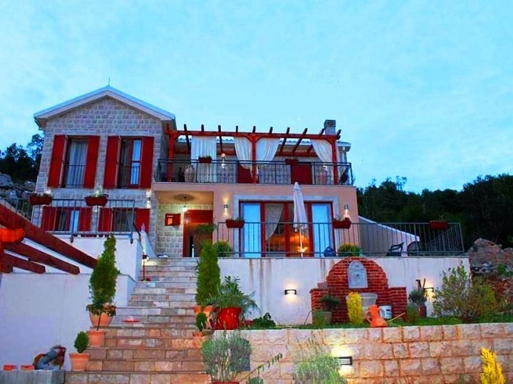 Two-story stone villa in Krimovica, Montenegro, with a terrace, red shutters, and landscaped grounds.