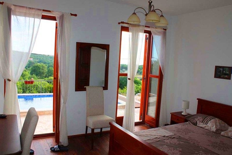 Bedroom with a double bed and access to the pool at a villa in Krimovica, Montenegro, overlooking a green garden and terrace.