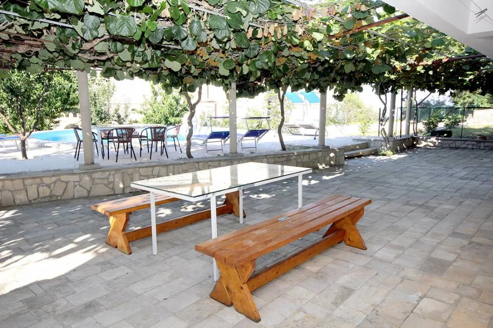 Open patio with wooden benches and glass table under a leafy pergola, pool visible in the background.