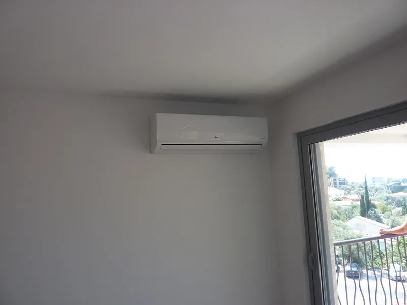 Empty white wall with an air conditioner and a glass door leading to a balcony showing outdoor greenery.