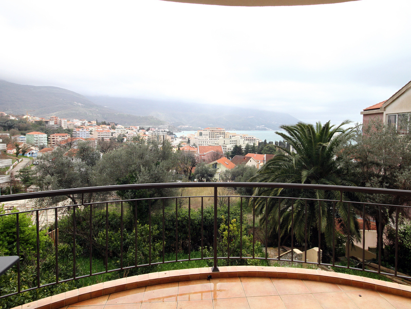View from the apartment balcony in Bečići, Montenegro: panoramic view of the city, mountains, and sea, tiled flooring, wrought-iron railing.