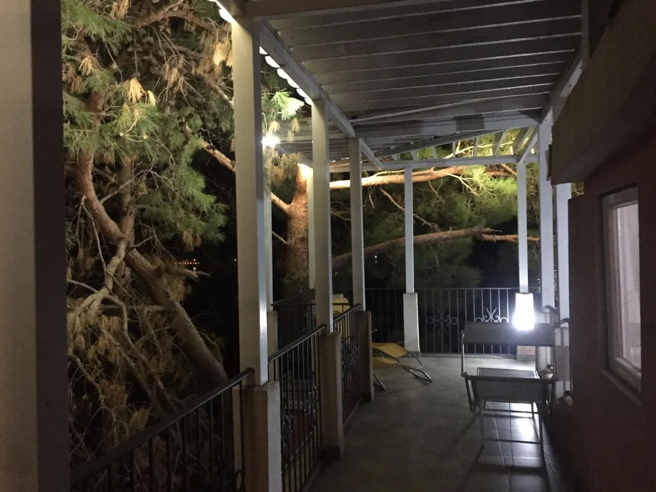 Dark balcony with metal railing, columns, and roof; pine trees visible outside along with folding chairs.