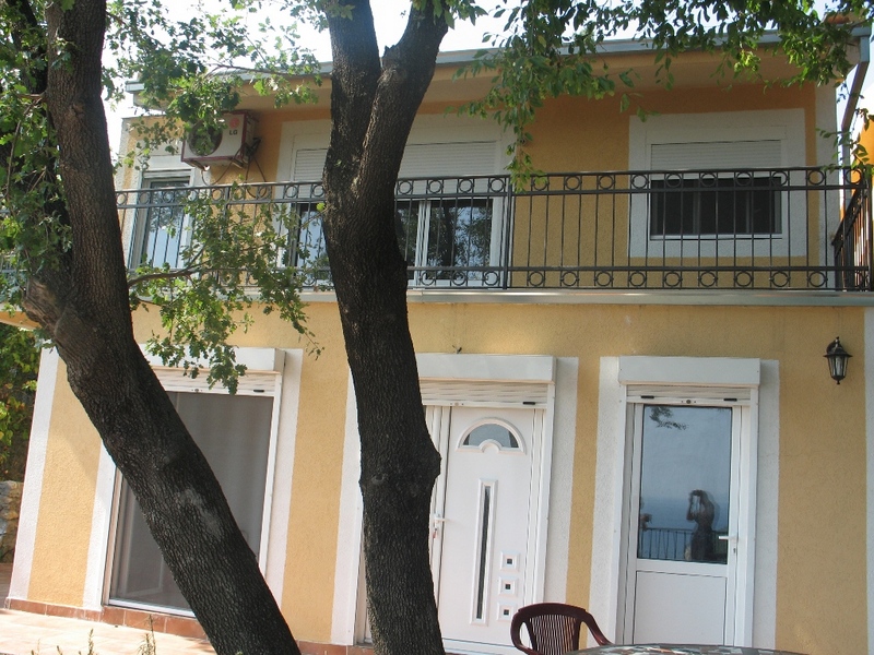 Two-story house in Sutomore, Montenegro: facade with a balcony, wrought-iron railing, and a terrace surrounded by trees.