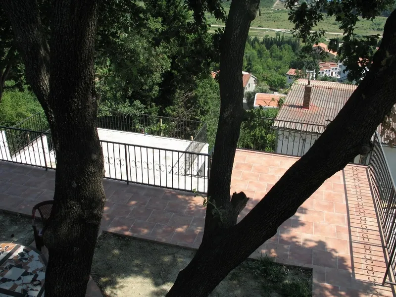Terrace with terracotta tiles and black metal railing, overlooking trees and neighboring houses.