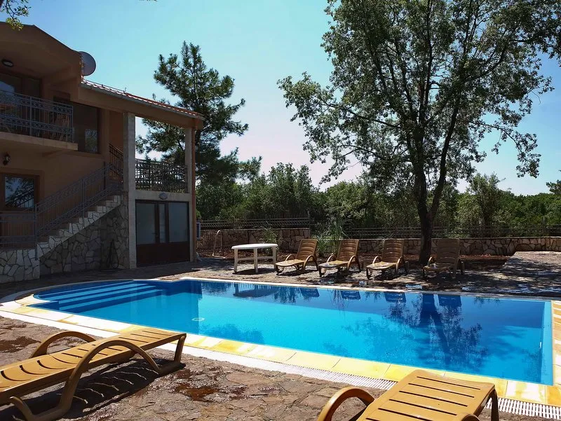 Outdoor swimming pool with blue water and wooden sun loungers on a stone patio next to a house.