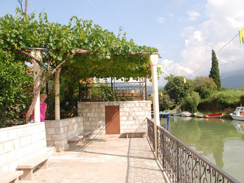 Terrace with grapevines and stone benches by the river in Igalo, Montenegro, view of the pier with boats.