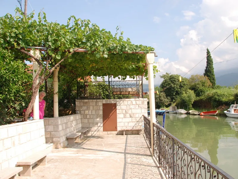 Open stone terrace with a grapevine pergola and wrought iron railing overlooking water with boats.
