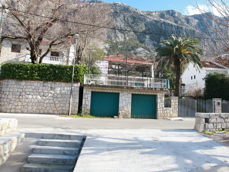 House in Dobrota, Montenegro: facade with a garage, stone detailing, terrace, and mountain views surrounded by palm trees.