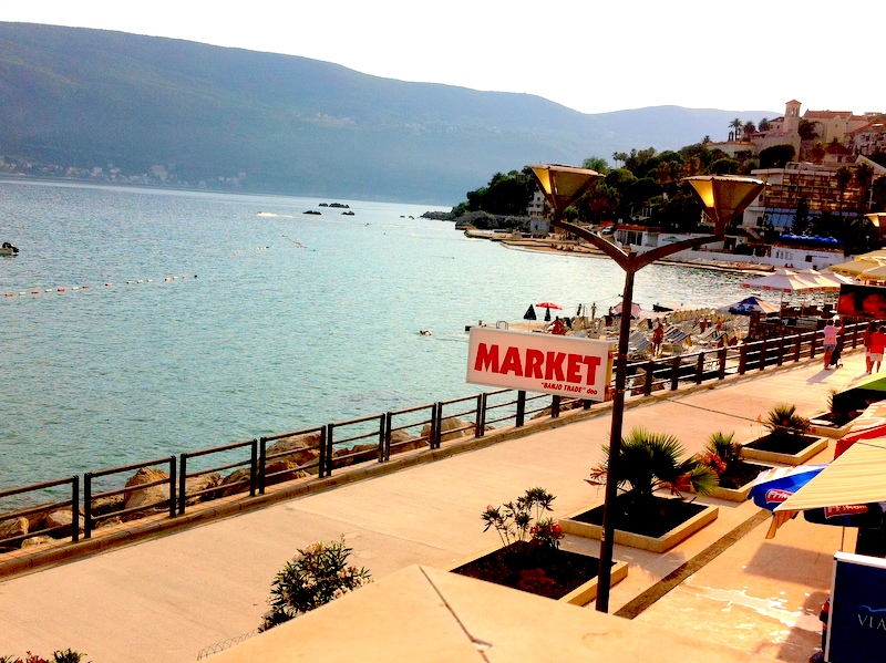 View of the waterfront, beach, and sea in Herceg Novi, Montenegro: a promenade with palm trees and a shop by the coast.