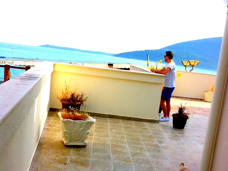 Tiled terrace overlooking the sea with white walls, potted plants, and a man looking at the water view.