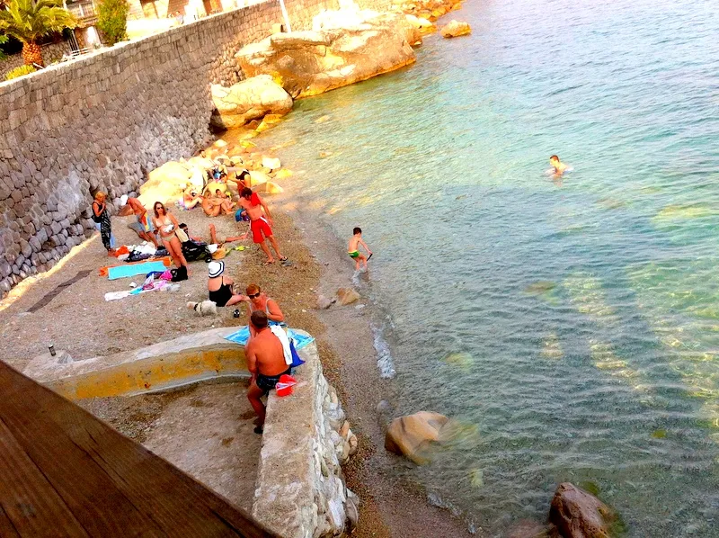 People sunbathing and swimming at a rocky beach next to a stone wall and sea.