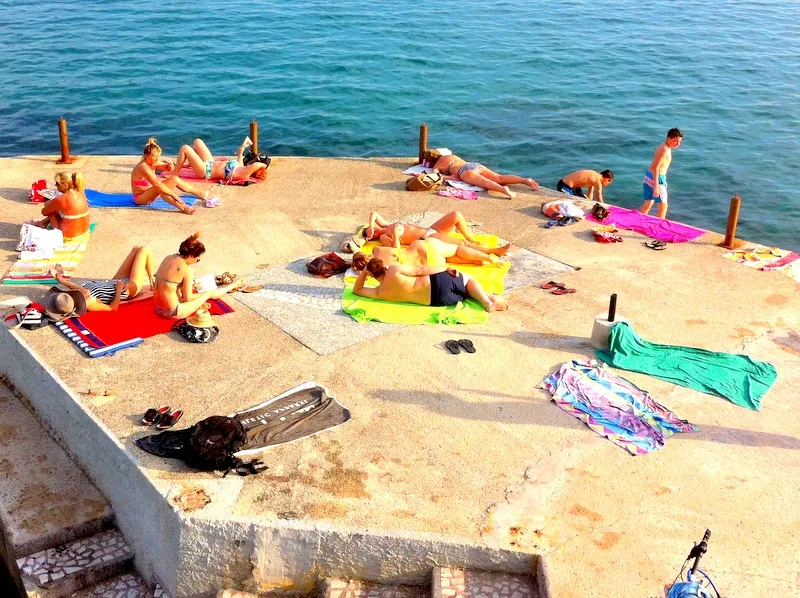 People in swimwear sunbathing on a concrete pier by the sea with towels and personal items scattered around.