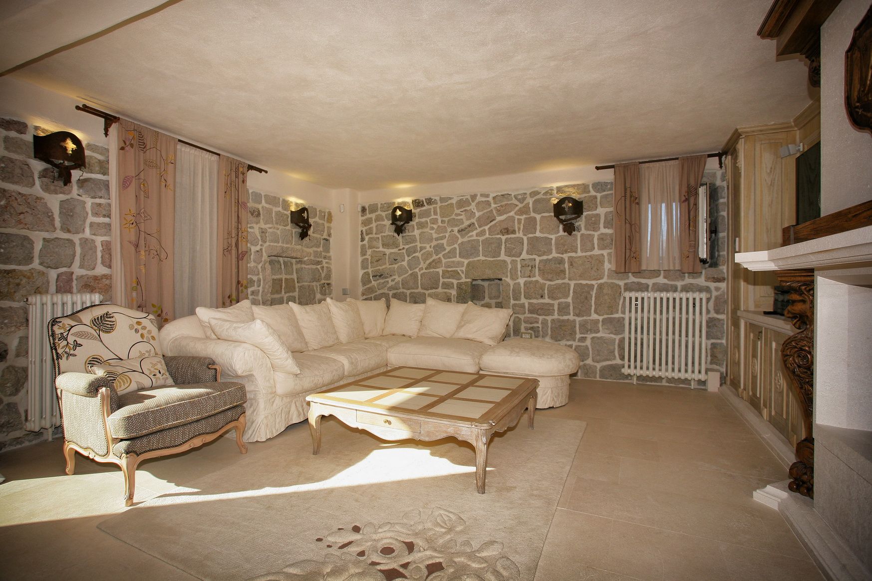 Villa living room in Reževići, Montenegro: stone walls, a light corner sofa, a carved fireplace, and classic furniture.