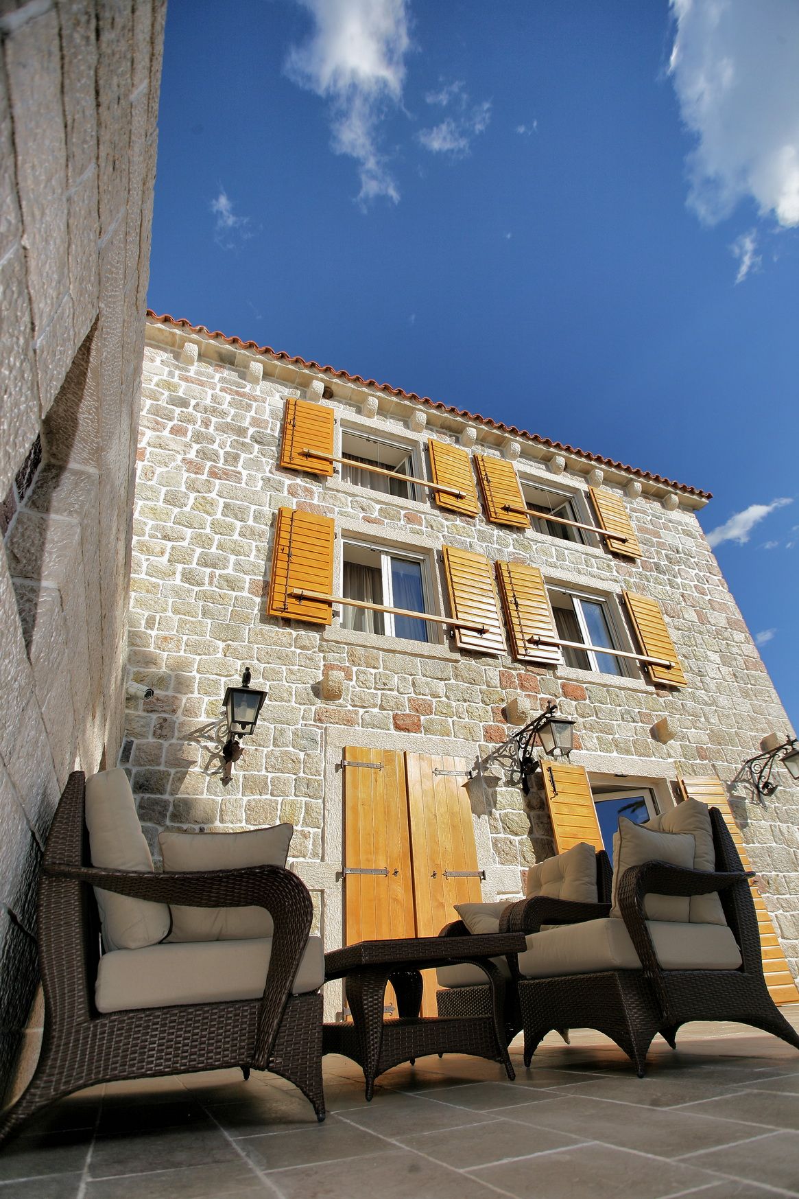Facade of a stone villa with wooden shutters and a terrace with wicker furniture in Reževići, Montenegro.