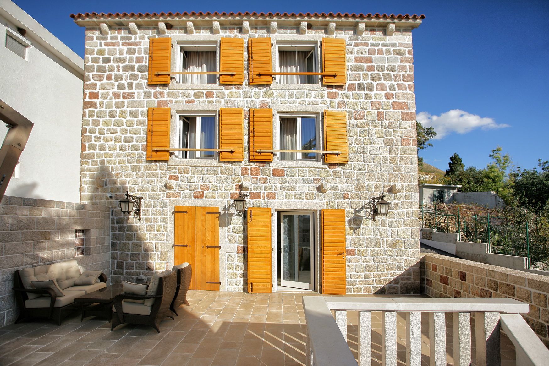 Three-story stone villa in Reževići, Montenegro, with a terrace, wicker furniture, and wooden shutters on the windows.