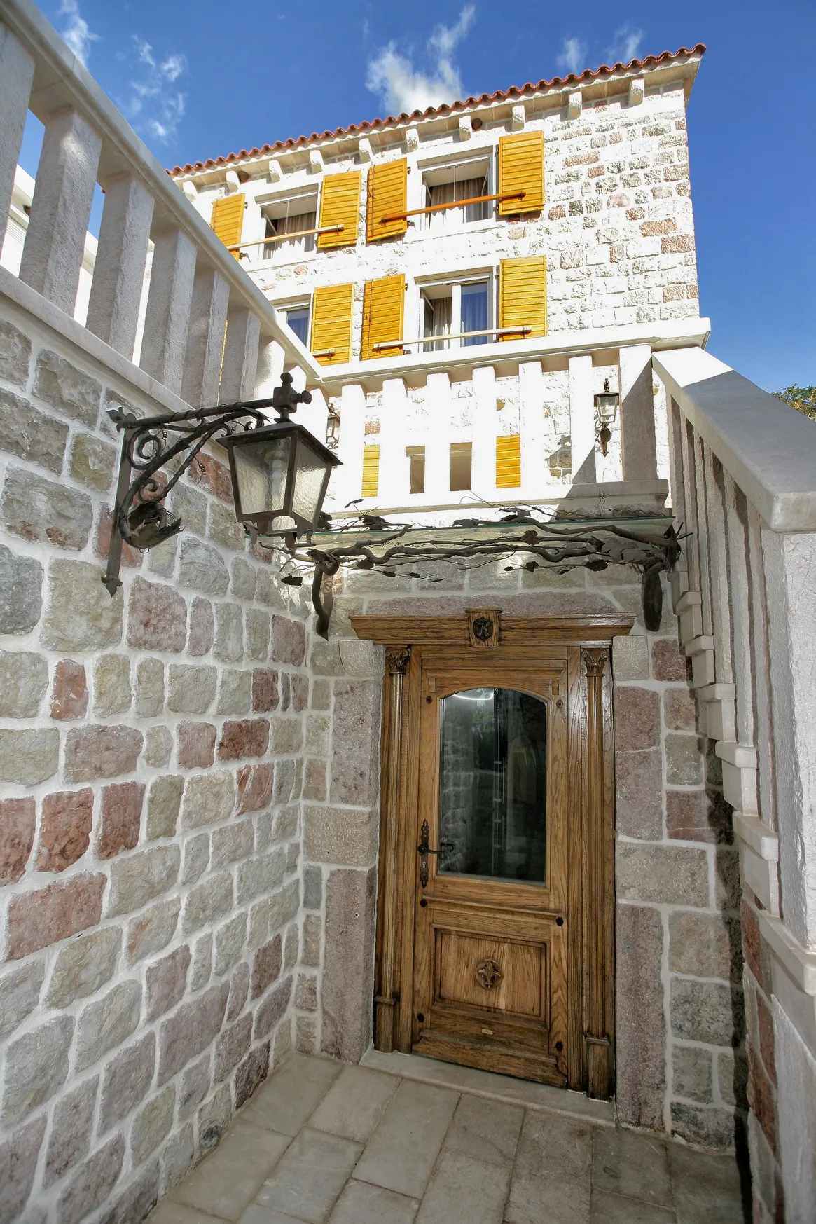 Stone entrance with a wooden door, wrought iron lantern, and yellow shutters under a blue sky.
