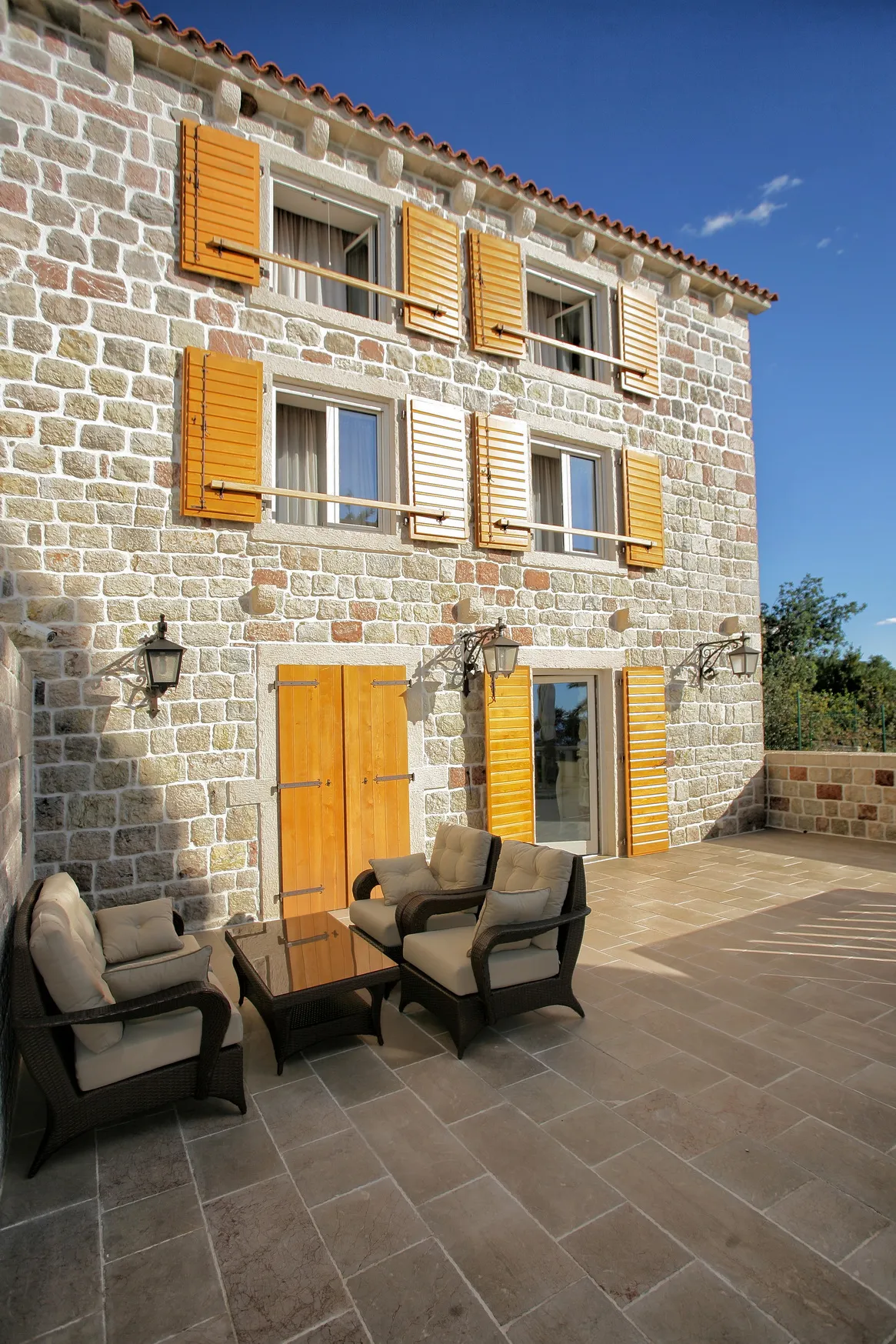 Stone house facade with wooden shutters and outdoor furniture on a tiled terrace under a clear blue sky.