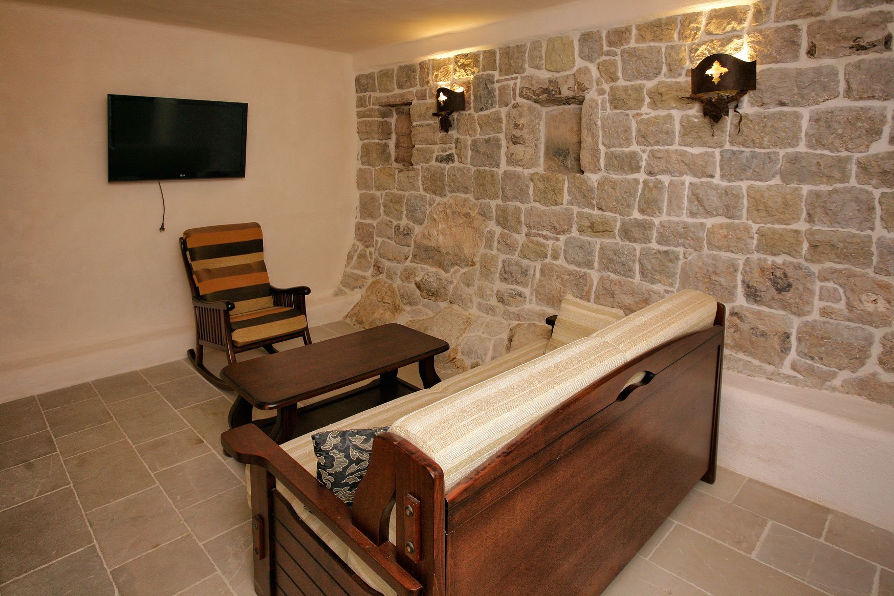Living room with a stone wall and wooden furniture in a villa in Reževići, Montenegro. Cozy interior with a TV and an armchair.