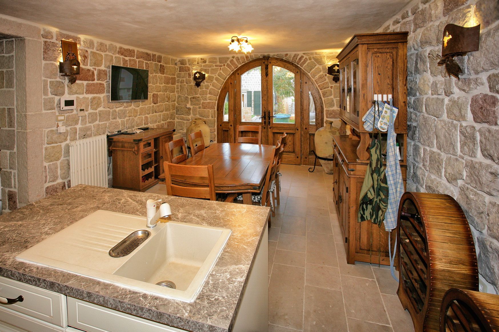 Kitchen-dining room with stone walls and wooden furniture in a villa in Reževići, Montenegro. Arched door and courtyard view.