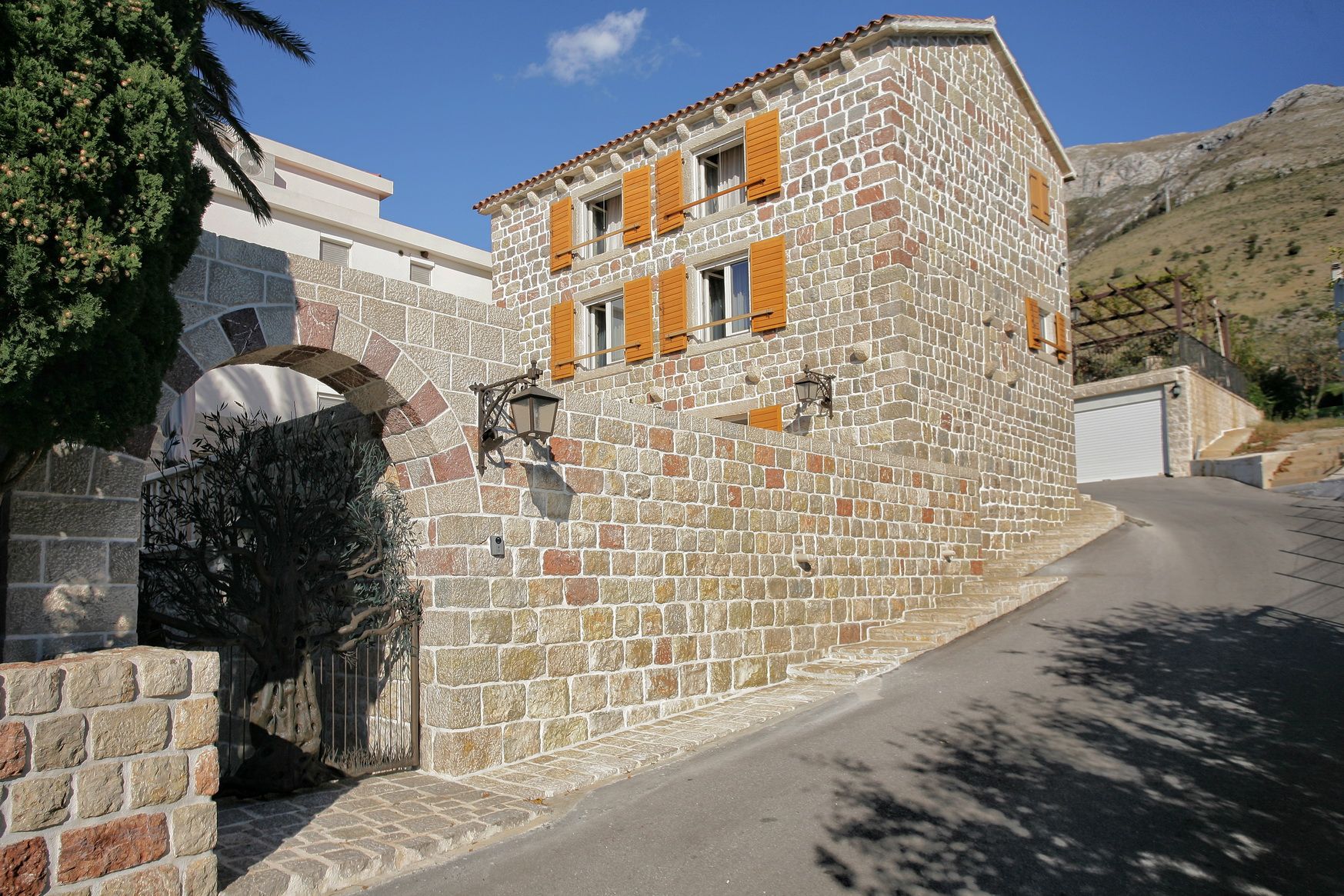 Traditional stone villa with yellow shutters in Reževići, Montenegro: front view and arched entrance from the street.