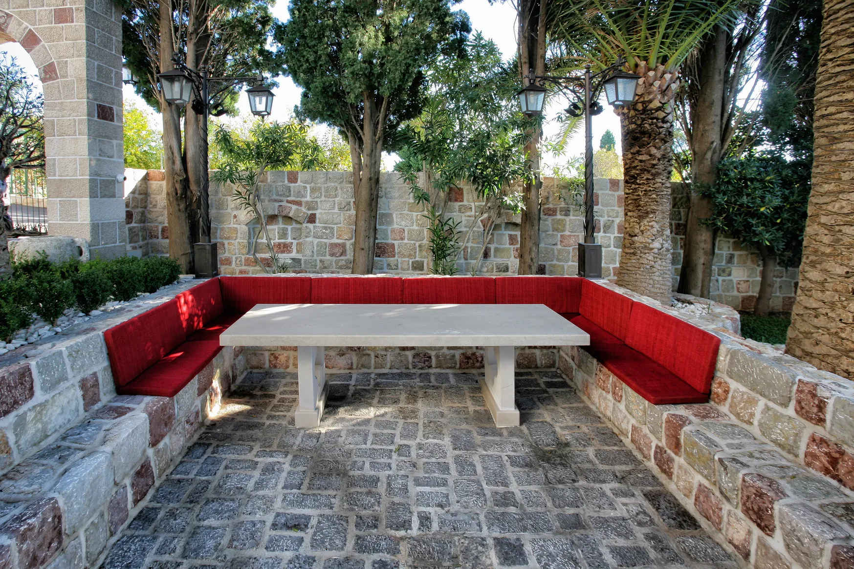 Outdoor stone patio with red cushioned benches and a stone table surrounded by trees and lanterns.