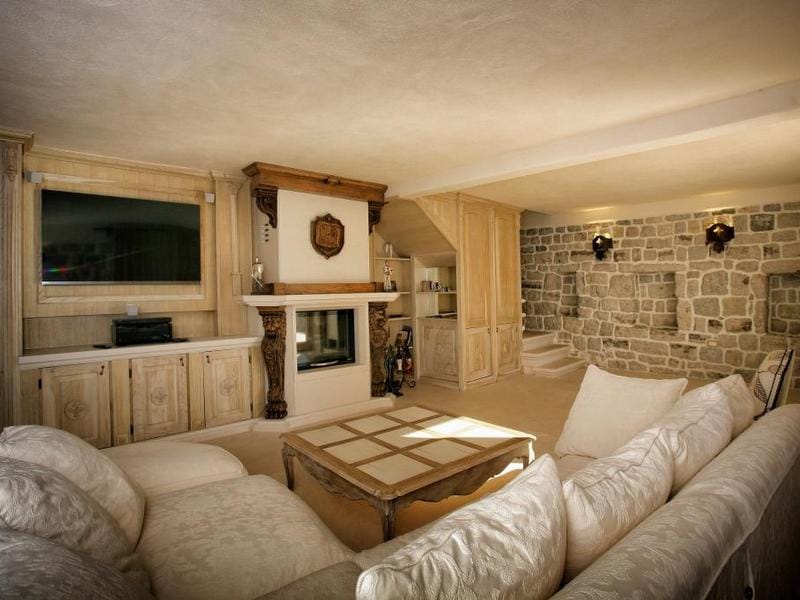 Villa living room in Rezevici, Montenegro: stone walls, a fireplace with carved decor, light furniture, and a wooden TV area.