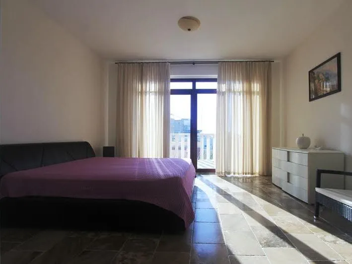 Bedroom with a purple bedspread, white dresser, and balcony visible through beige curtains.