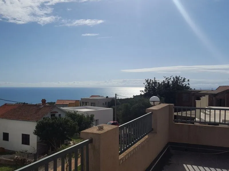 View from a balcony overlooking the ocean and neighboring houses with red roofs under a clear blue sky.