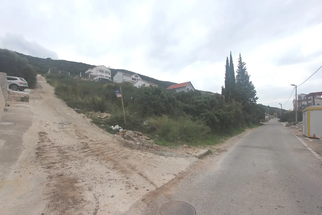 View of a dirt and asphalt road intersection with houses on a hill, trees, and an overcast sky in the background.