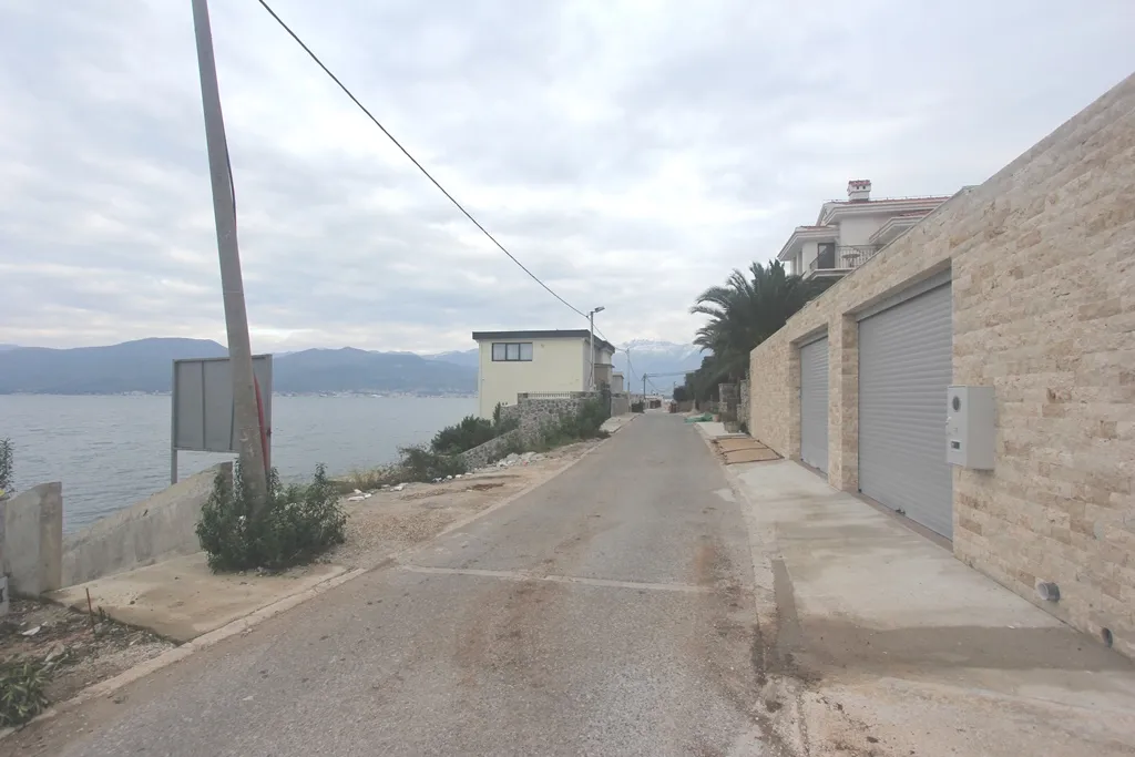 Paved road along the water featuring a stone wall and garage doors under an overcast sky.