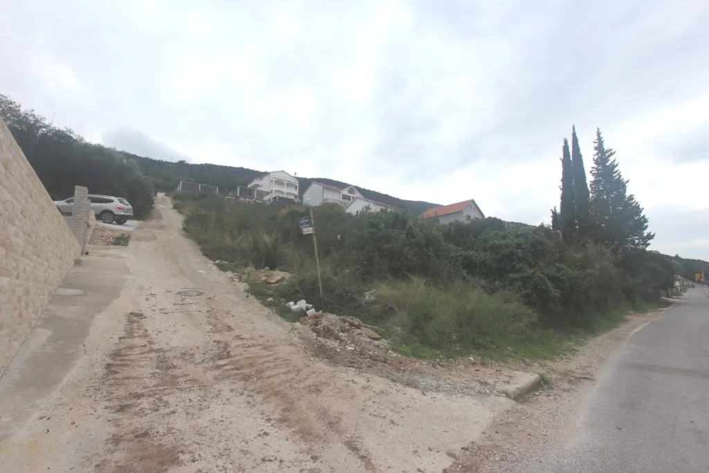 Exterior view: dirt road, stone wall, vegetation, and houses on a hill under a cloudy sky.