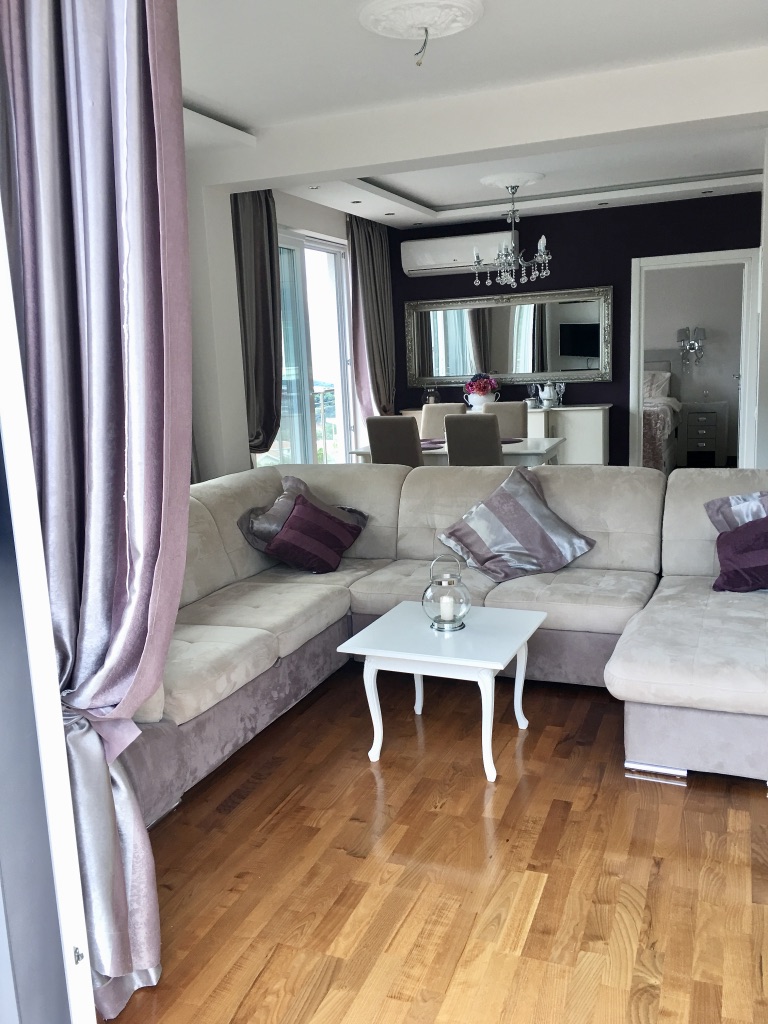 Living room with an L-shaped sofa, dining area, and parquet flooring in an apartment in Bečići, Montenegro. Modern interior.
