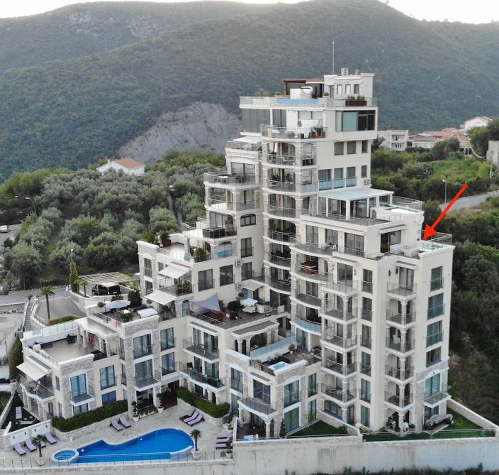 Aerial view of a residential complex in Bečići, Montenegro: a multi-level building with terraces, a pool, and mountains in the background.