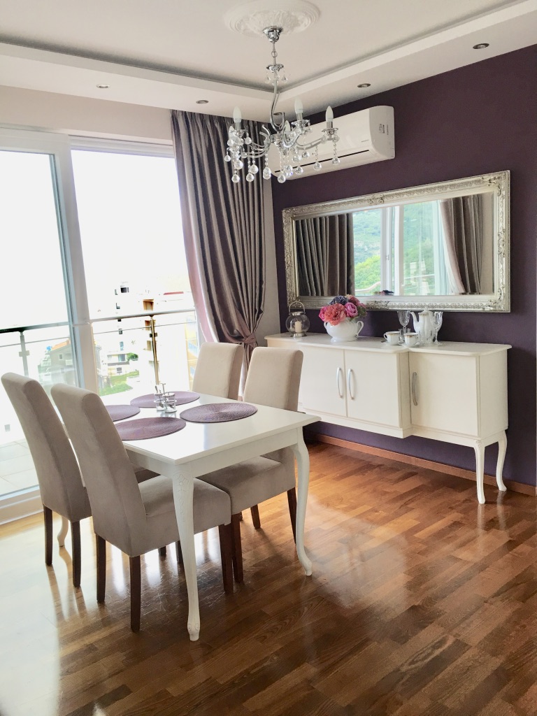 Bright dining area in an apartment in Bečići, Montenegro: white furniture, parquet flooring, a purple wall, and a panoramic window.