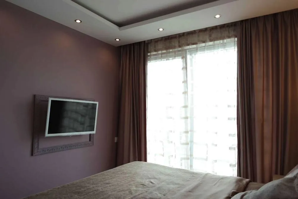 Bedroom with purple wall, framed TV, and bed near a window with brown curtains.