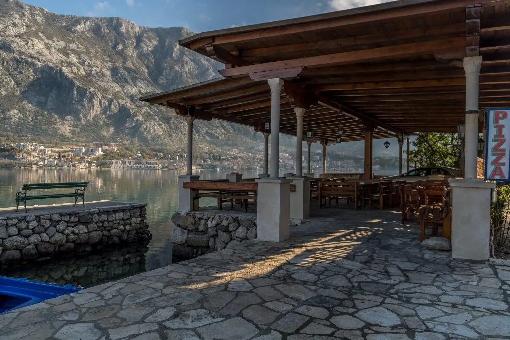 Open stone terrace with wooden roof and furniture by the water against a mountain backdrop.