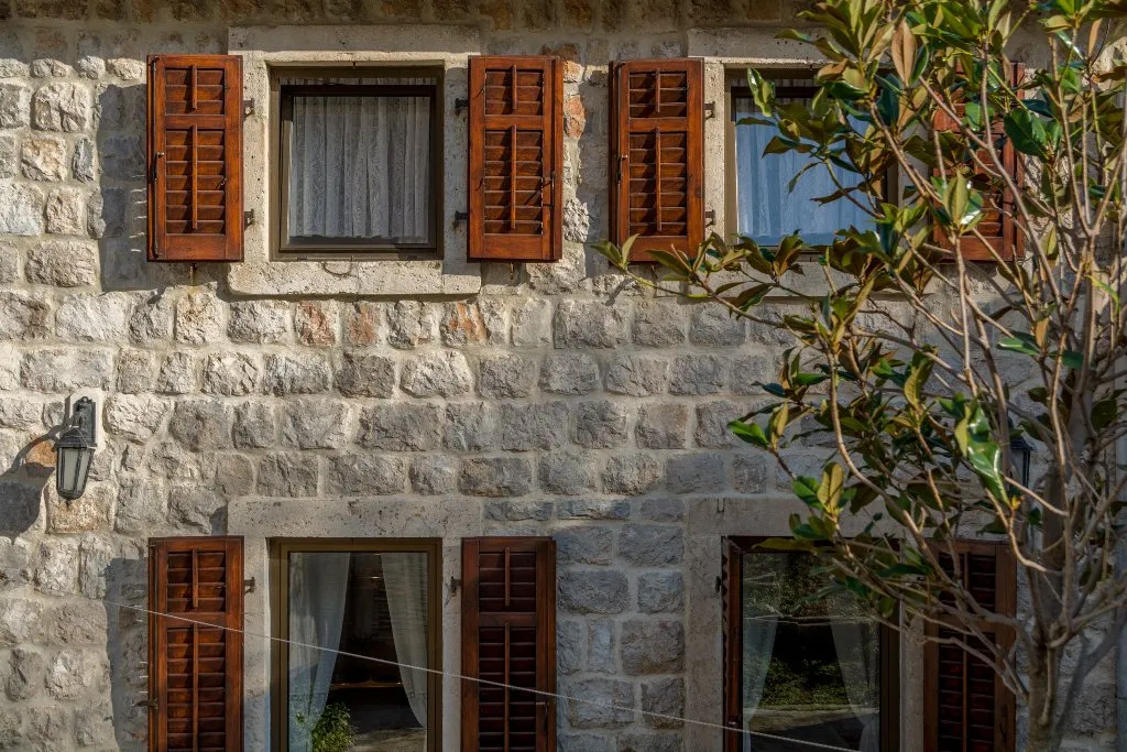Stone building facade featuring wooden window frames and shutters, with tree branches visible on the right side.