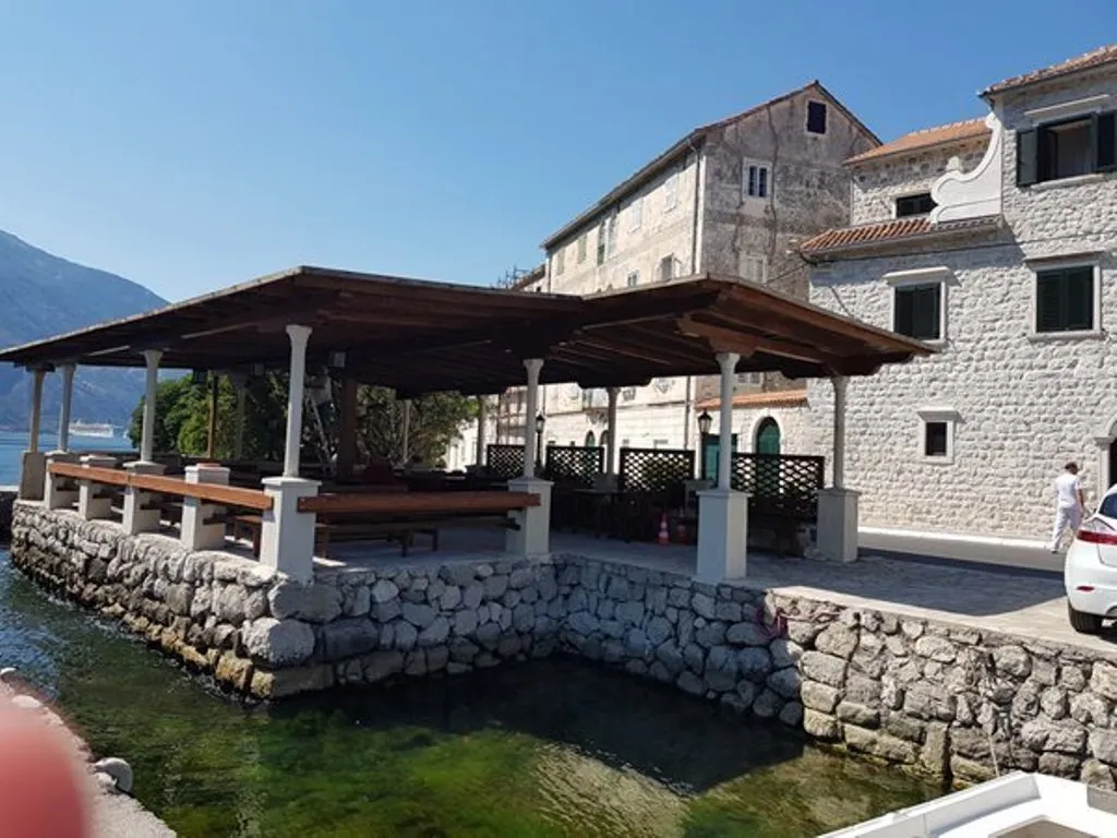 Open stone terrace with wooden pergola by the water, adjacent to a historic stone building and mountains.