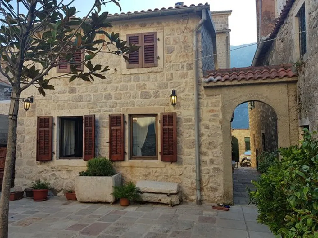 Stone house facade with wooden shutters, an arched passageway, potted plants, and a tree on a paved courtyard.
