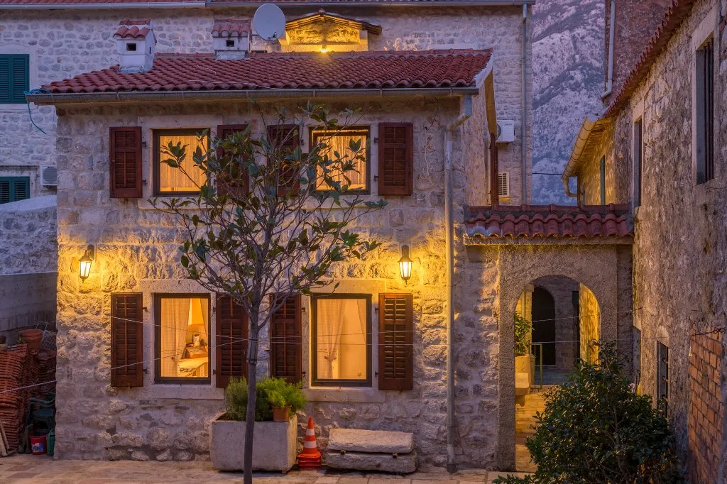 Exterior of a stone house with red tile roof, wooden shutters, and warm lit windows at dusk.